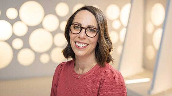 Nina Lopez-Corrado smiles at the camera. She is wearing brown glasses and a red sweater and standing in front of a white backdrop.