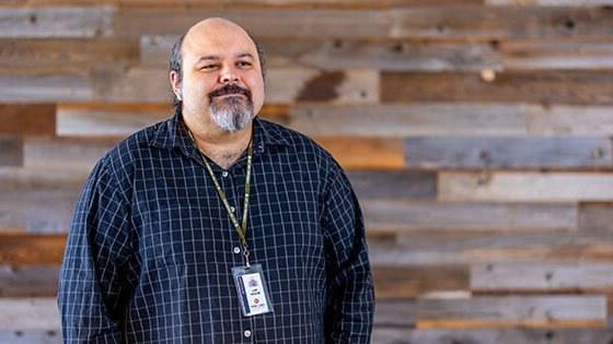 Instructor Eric Berzins, a man with a grey goatee, standing in front of a wood paneled wall wearing a blue button down dress shirt and green Full Sail lanyard with name badge.