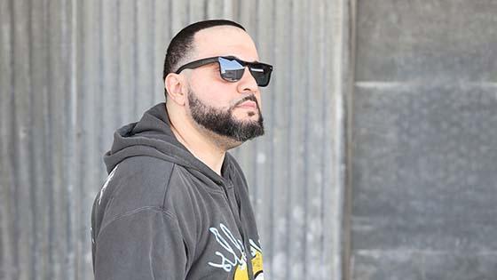 满帆 grad George Doman wearing black sunglasses and a grey pullover hoodie with a yellow and light blue design, standing in front of a large stone wall while looking off into the distance.