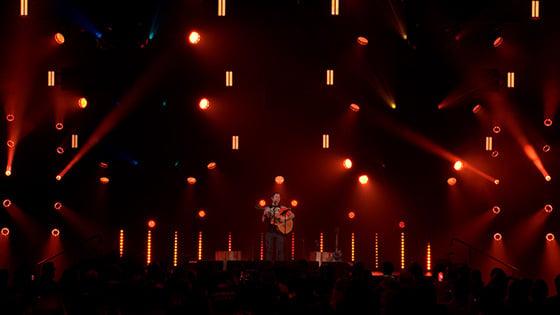 Trivium lead-singer Matt Heafy on a dimly-lit stage playing an acoustic guitar in front of a large crowd.