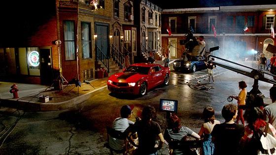 Full Sail University's Backlot in the evening filled with a full film production crew shooting a scene featuring two muscle cars, one red and one black.