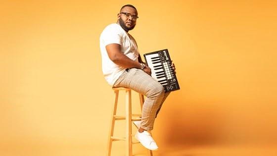 Chavez Parker sits on a stool and holds a keyboard. He wears glasses, a t-shirt, brown pants, and sneakers.