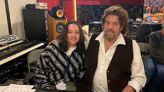 Alan Parsons sits with Steffie Tjandra in front of a mixing board in a recording studio. They are smiling at the camera.