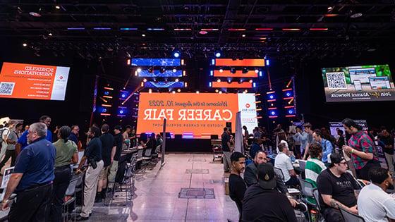 A bustling career fair with several booths set up. In the center, a giant LED screen welcomes guests to the Career Fair.