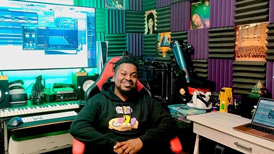 Grad Shawn Deshield smiling while seated at a desk in a home studio featuring a keyboard, audio equipment, and a large screen displaying audio software while wearing a black hooded sweatshirt with a colorful design.