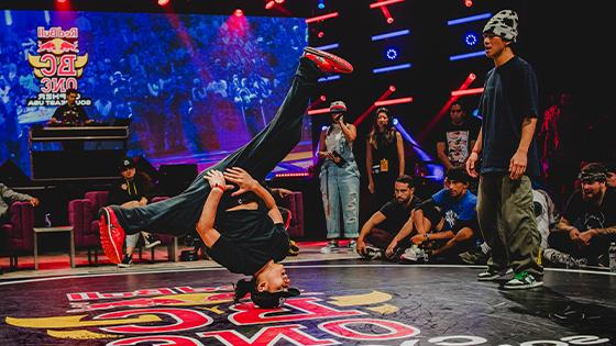 A dancer is doing a hands-free headstand with their legs spread while their competitor, the audience, and the judges watch.