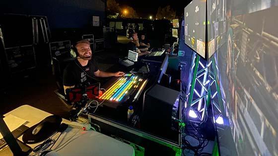 A smiling young man wearing headphones and sitting at a video switcher looks up at two large video screens in a production studio.