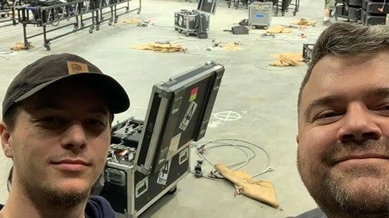 Two smiling men with beards stand in a backstage area. There are wires on the floor behind them, as well as an open trunk filled with production equipment.