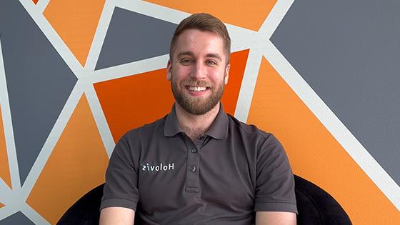 A man with light hair and a beard wearing a 灰色 Holovis polo while seated and smiling in front of an orange, 灰色, and white geometric background.