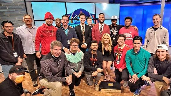 A group shot of several Full Sail students and instructors in front of a large screen on set at the Dan Patrick School of Sportscasting.