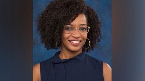 Tiana, an African American woman, sits smiling at the camera. She is wearing a sleeveless navy blue blouse, silver hoop earrings, and glasses with translucent beige frames.