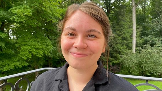 Emmi Litsenberger sits on a porch in front of a grassy lawn and trees. 她在微笑 and wearing a gray button-up shirt.