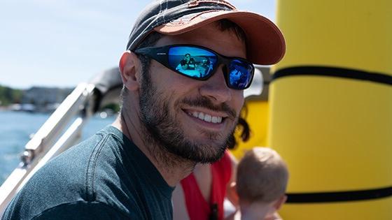 一个穿蓝色t恤的男人, 一顶蓝红相间的棒球帽, and sunglasses with polarized lenses sits on a boat on a sunny day. He is smiling at the camera and there is a brunette woman and a blonde child sitting out of focus behind him.