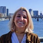 Grad Angela Decastro, a woman with shoulder-length blonde hair wearing a brown jacket and white top, smiles while standing on a bridge overlooking a river, in the distance behind her is a bridge and cityscape.