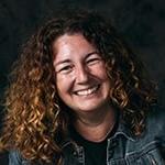 A woman with curly auburn hair smiling while wearing a black tee and denim jacket against a black backdrop.