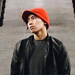 A young Asian man wearing a red knit cap, black jacket and shirt, green pants, and a burgundy bag stands between two escalators. His hands are in his pockets and he is looking off to the left.