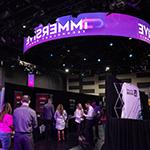 A large room with lots of booths set up, each from a different technology company. People in business-casual clothing are walking around and socializing.