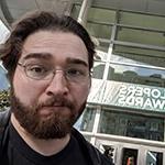 Ryan Miller pictured from above the shoulders in a black leather jacket with silver wireframe glasses in front of a sign reading “Game Developers Choice Awards.”