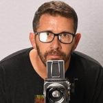 Sebastian Krys crouches behind a video camera. He is wearing black-rimmed eyeglasses and a black t-shirt with a logo on it.