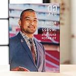 A 平面设计 USA magazine stands upright on a desk in a backlit room. 封面上, a man wearing a blue suit with a pink ribbon pin and holding an NFL football smiles for the camera. The cover reads, People to Watch in 2022, 学生 to Watch.