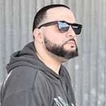 Full Sail grad George Doman wearing black sunglasses and a grey pullover hoodie with a yellow and light blue design, standing in front of a large stone wall while looking off into the distance.