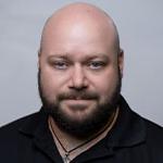 Grad Corey Guerra is wearing a black polo shirt and posing for a headshot against a light grey backdrop.