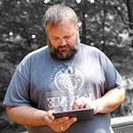 Grad Ryan Backa in a blue shirt with a white dragon design, holding an electronic tablet in front of a wooded area in greyscale.