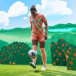 An Orlando Pride player kicks a soccer ball toward a camera on a film set that features an animated orange grove.