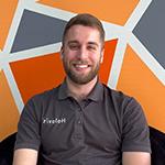 A man with light hair and a beard wearing a grey Holovis polo while seated and smiling in front of an orange, grey, and white geometric background.