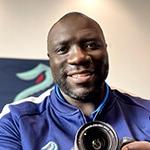 Grad Lamont Buford sports a blue jacket with a Seattle Kraken logo while smiling and holding a camera.