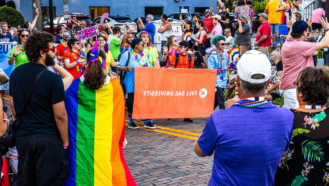 A group of people walking down the street through a parade carrying a large orange banner that reads “Full Sail University.”