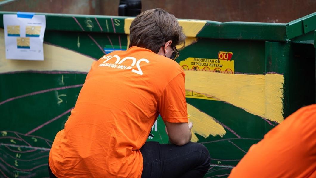 Man wearing a bright orange t-shirt crouches down with his back to the camera as he paints a large green trashcan.