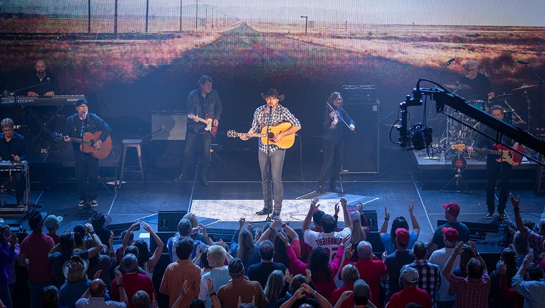 A man wearing a cowboy hat and flannel shirt is seen center stage while playing the guitar with an ensemble band in the background.