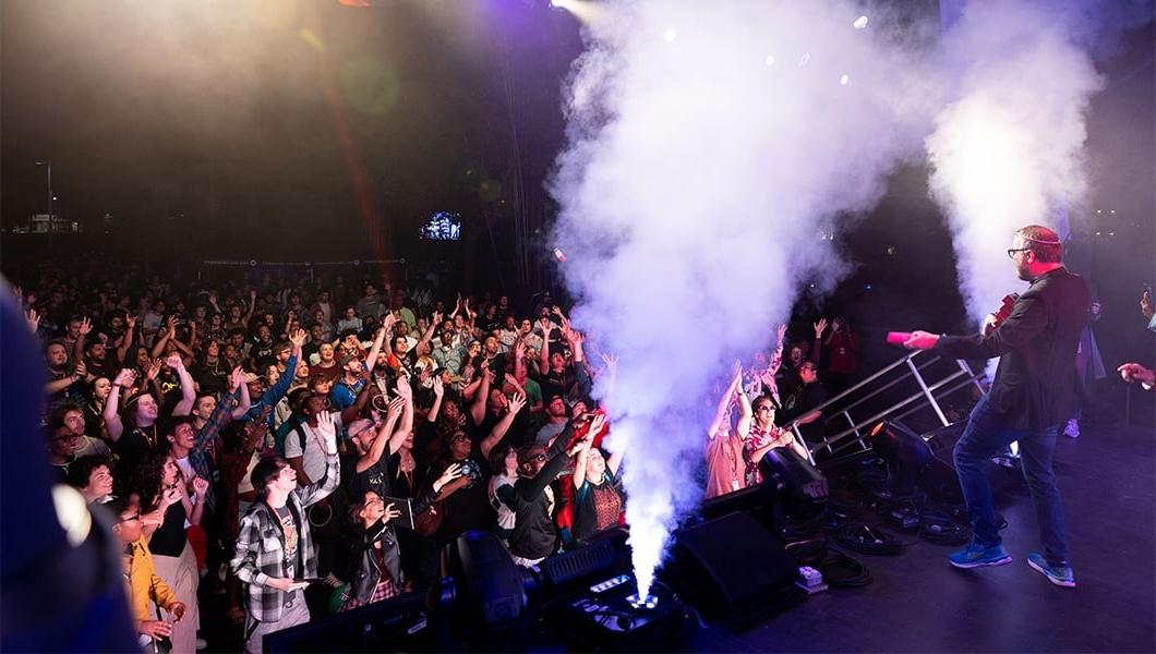 A man in a black blazer is throwing prizes into the crowd. There are two smoke machines on stage. 观众们面带微笑，他们伸向舞台前面，想要抓住奖品.